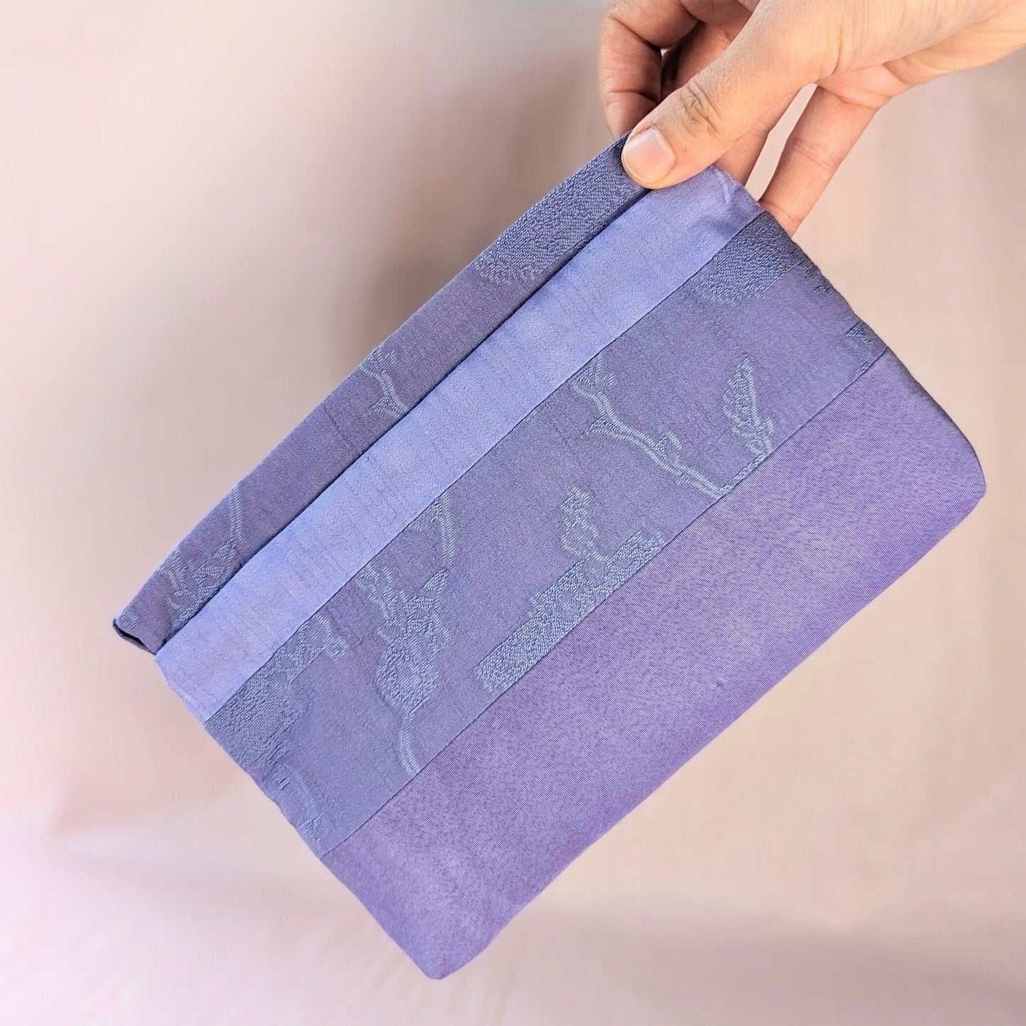 Image shows a white hand holding a lavender coloured, rectangle shaped purse. It is made of unequal stripes of fabric. Each fabric is a similar shade of purple but each is a different texture. The background is pale pink.