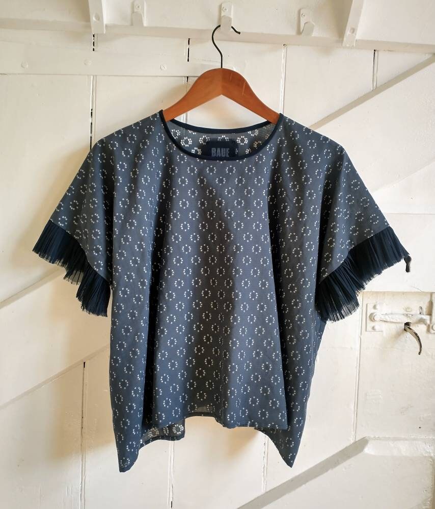 The Frida top shown on a hanger. The blouse falls lower at the sides than the centre front so it creates a gently curved hem when on the body. A navy blue brand label is visible on the inside of the back neck with Baue printed in light blue.