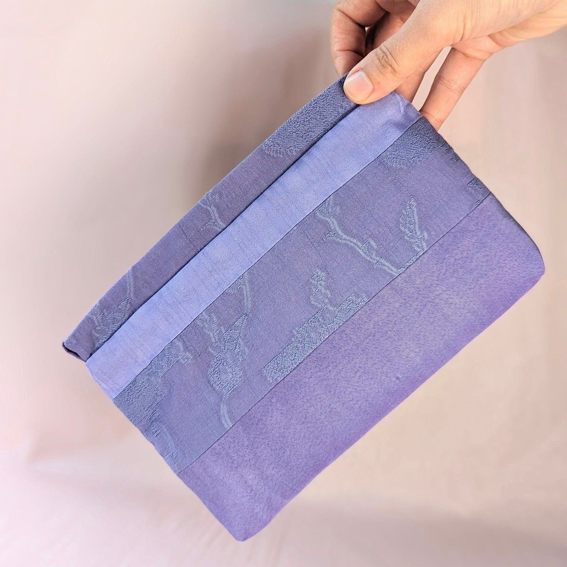 Image shows a white hand holding a lavender coloured, rectangle shaped purse. It is made of unequal stripes of fabric. Each fabric is a similar shade of purple but each is a different texture. The background is pale pink.