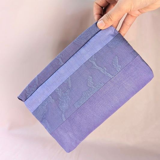 Image shows a white hand holding a lavender coloured, rectangle shaped purse. It is made of unequal stripes of fabric. Each fabric is a similar shade of purple but each is a different texture. The background is pale pink.